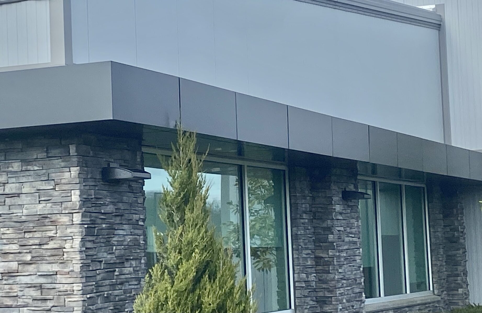 Modern building facade with stone and metal panel design, featuring large windows and a small coniferous tree in front.