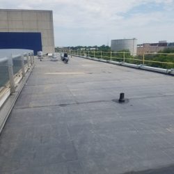 Flat rooftop with protective railings, pipes, and vents, surrounded by a mix of industrial and natural landscape. Cloudy sky above.
