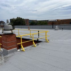 A flat rooftop with yellow safety railings, a chimney, and two ventilation units under a cloudy sky.