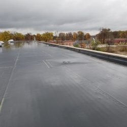 Flat rooftop with a drain and HVAC units under a cloudy sky, surrounded by trees and buildings in the distance.