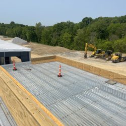 Construction site with metal roofing, wooden barriers, orange cones, and construction vehicles in a wooded area.