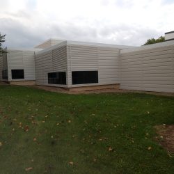 Modern white metal building with multiple sections, surrounded by a grassy area and some fallen leaves. Overcast sky.